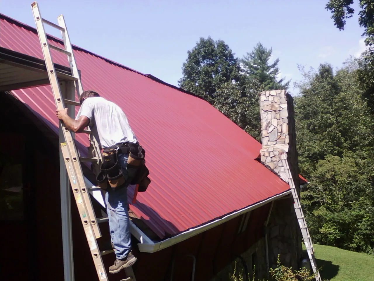 Roofer on a ladder beside a red metal roof and stone chimney — Soffit & Fascia Repair by Roofing Brothers in Goodells